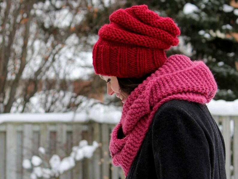 May include: A bright red chunky knit beanie with a ribbed brim and a matching deep pink chunky knit infinity scarf worn over a dark coat. The accessories feature a thick, textured yarn construction designed for cold weather wear.