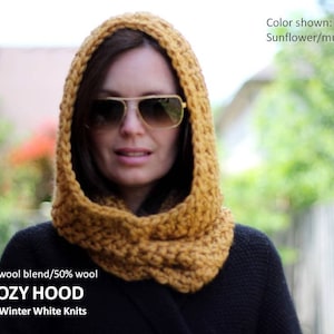 May include: A mustard yellow knitted hooded cowl made from a soft wool blend. The cowl is worn by a person with brown hair and sunglasses. The text "COZY HOOD" and "By, Winter White Knits" is visible in the image.