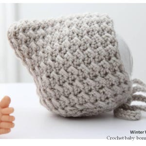 May include: A light grey, textured crochet baby bonnet with tie strings. The bonnet is displayed on a white surface. The text "Winter White Knits" and "Crochet baby bonnet pattern" are visible.