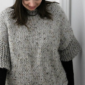 May include: A gray knitted sweater with a speckled pattern. The sweater has a round neckline and long sleeves. The text "KNITTING PATTERN Winter White Knits" is visible in the top right corner of the image.