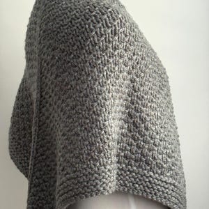 May include: A grey knitted shawl featuring a textured basketweave pattern throughout the main body. The garment has a smooth, garter stitch border along the edges, providing a structured finish to the soft, thick yarn construction of the piece.