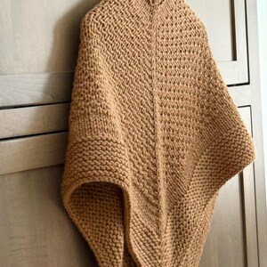 May include: A tan knitted poncho featuring a textured stitch pattern with a central vertical seam. The garment has a high collar and a triangular silhouette, crafted from thick yarn with a consistent light brown colour throughout the piece.