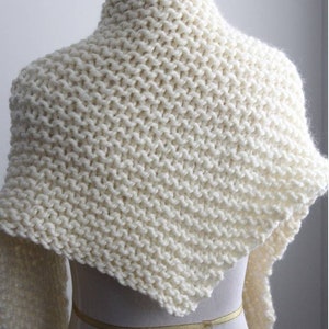 May include: A chunky knit shawl in an off-white colour featuring a garter stitch pattern draped over a mannequin. The shawl has a triangular shape with a high neck collar. The text "Winter White Knits" is printed in white at the bottom centre.