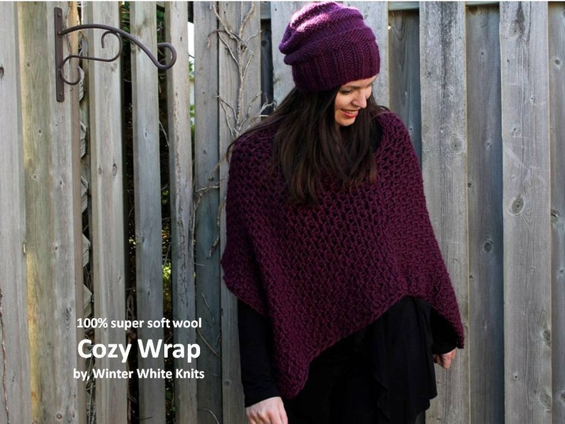 May include: A purple knitted wrap made from 100% super soft wool. The wrap is worn by a person standing in front of a wooden fence. The text "Cozy Wrap" and "by Winter White Knits" is printed below the wrap.