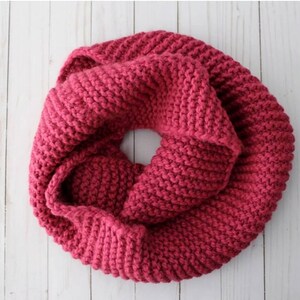 May include: A chunky knit infinity scarf in a deep magenta color. The scarf features a thick, textured garter stitch pattern and is arranged in a circular, folded loop shape, showcasing the dense, soft yarn construction throughout the piece.