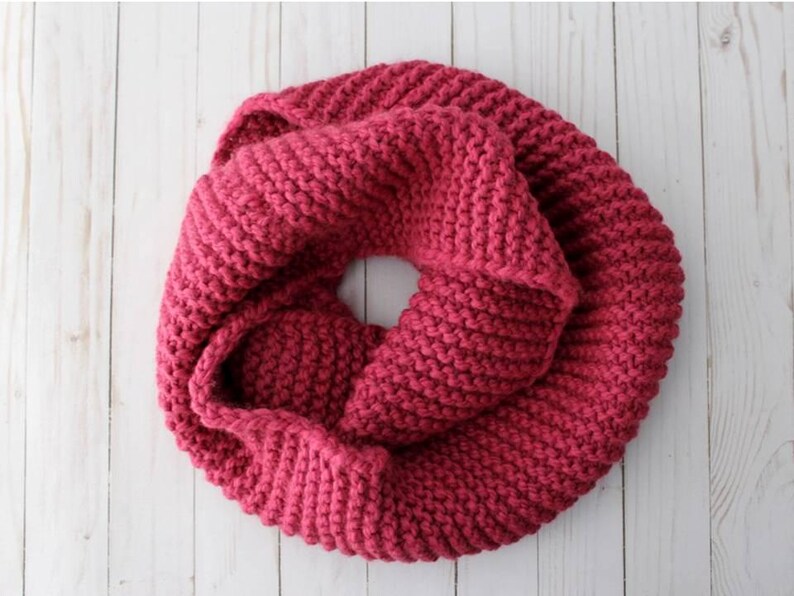 May include: A chunky knit infinity scarf in a deep magenta color. The scarf features a thick, textured garter stitch pattern and is arranged in a circular, folded loop shape, showcasing the dense, soft yarn construction throughout the piece.