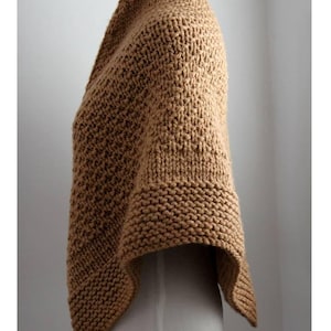 May include: A tan knitted poncho featuring a textured stitch pattern throughout the body and a distinct garter stitch border along the hem. The garment is displayed on a mannequin, showcasing its draped silhouette and thick yarn construction.