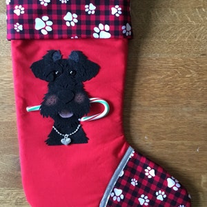 May include: Red Christmas stocking with a black and white buffalo plaid pattern and white paw prints. A black felt dog with a silver heart charm is on the stocking. A candy cane is in the dog's mouth.