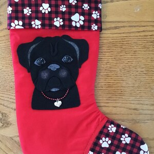 May include: Red Christmas stocking with a black pug face appliqué. The stocking has a black and red buffalo plaid cuff with white paw prints.