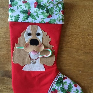 May include: Red Christmas stocking with a felt applique of a brown and white dog with a candy cane in its mouth. The stocking is decorated with a holly pattern.