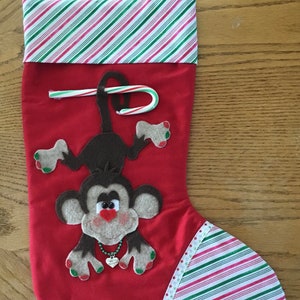 May include: Red Christmas stocking with a green and white striped cuff. A brown felt monkey with green nail polish is hanging from the stocking. A candy cane is tucked into the stocking.