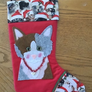 May include: Red Christmas stocking with a felt cat face appliqué. The cat has brown, white, and gray fur, green eyes, and a red and white beaded necklace with a heart charm. The stocking is decorated with a pattern of cats wearing Santa hats.