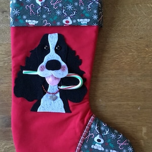 May include: Red Christmas stocking with a green and white patterned cuff. The stocking features a felt applique of a black and white dog with a candy cane in its mouth.
