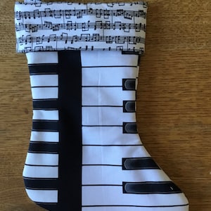 May include: A white and black Christmas stocking with a piano keyboard design. The stocking has a loop at the top for hanging and is made of fabric.