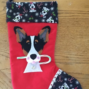 May include: Red Christmas stocking with a black and white dog face applique. The dog is holding a candy cane in its mouth and wearing a silver heart charm necklace. The stocking is trimmed with black fabric with a repeating pattern of white dogs wearing Santa hats.