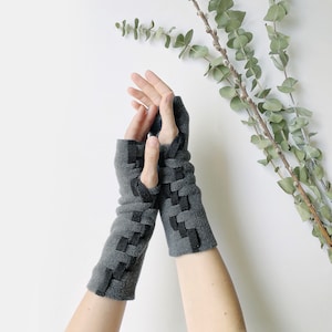 May include: Gray fingerless gloves with a woven black design. The gloves are made of a soft material and are designed to keep hands warm while allowing for finger movement. A stylish accessory for cold weather.