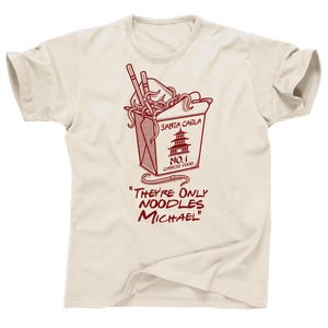 Op de afbeelding: Crèmekleurig T-shirt met een rode afbeelding van een Chinees voedselbox met noedels en eetstokjes. Op de doos staat "Santa Carla No. 1 Chinese Food". De tekst eronder luidt "They're Only Noodles Michael."