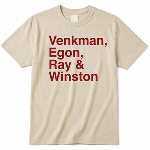 Op de afbeelding: Een beige t-shirt met korte mouwen en een ronde hals, voorzien van donkerrode tekst op de voorzijde. De tekst luidt: Venkman, Egon, Ray & Winston. Het kledingstuk heeft een standaard pasvorm met een geribbelde kraag en een rechte zoom.