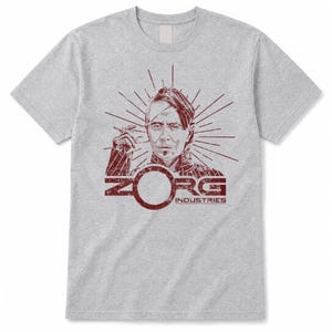 Pode incluir: Uma t-shirt de manga curta cinzenta clara com uma impressão gráfica vermelho-escura de uma pessoa com um penteado estilizado. O design inclui linhas radiantes atrás da figura e o texto ZORG INDUSTRIES com uma letra O circular.