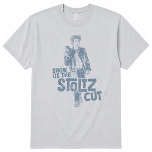 Puede incluir: Una camiseta gris claro de manga corta con un estampado gráfico azul de una persona con chaqueta que sostiene una cámara. El texto "SHOW US THE STOLTZ CUT" está impreso en azul debajo de la figura. El diseño tiene un aspecto desgastado.