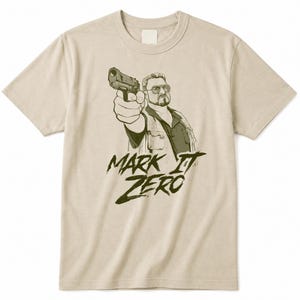 Może przedstawiać: Beżowy t-shirt z krótkim rękawem z ciemnozieloną grafiką przedstawiającą osobę w okularach i z bródką, celującą z pistoletu przed siebie. Poniżej ilustracji znajduje się napis "MARK IT ZERO" wykonany pogrubioną, ciemnozieloną czcionką.