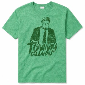 Peut inclure: Un t-shirt à manches courtes vert chiné avec une impression graphique vert foncé représentant un homme en costume et cravate, accompagné du texte Tommy Callahan imprimé en dessous. Le t-shirt présente un col rond et une coupe standard.