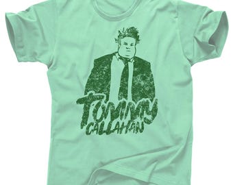 May include: A light green t-shirt featuring a green graphic of a person in a suit and tie. The text "Tommy Callahan" is printed below the graphic in a distressed font. The shirt has short sleeves and a crew neck.