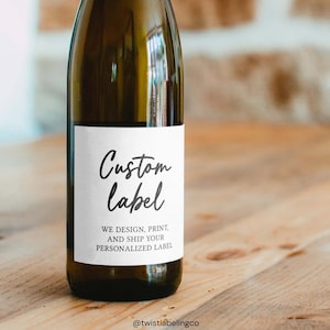 May include: A clear glass wine bottle with a white label. The label reads "Custom Label" in a handwritten font, with additional text below. The bottle sits on a wooden surface. The background includes a brick wall and more wood.
