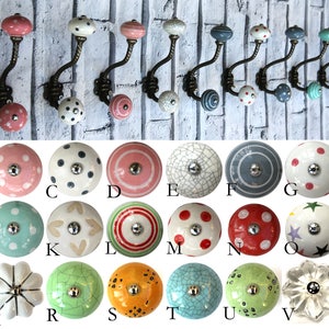 May include: An assortment of 26 ceramic knobs in various colours and patterns, including pink, green, blue, white, and red. The knobs feature polka dots, stripes, hearts, and other designs. The knobs are displayed on a white brick wall.