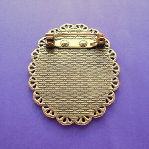 May include: An antique-style brooch with a decorative, oval-shaped frame. The brooch has a textured, bronze-colored surface and a scalloped edge. A pin and clasp are attached to the back for secure fastening. The brooch is set against a purple background.