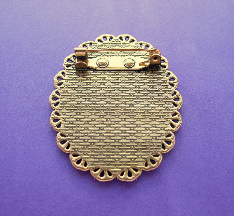 May include: An antique-style brooch with a decorative, oval-shaped frame. The brooch has a textured, bronze-colored surface and a scalloped edge. A pin and clasp are attached to the back for secure fastening. The brooch is set against a purple background.