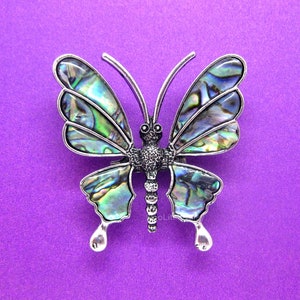 May include: A silver butterfly brooch with iridescent green and blue wings. The wings are made of abalone shell and have a textured surface.