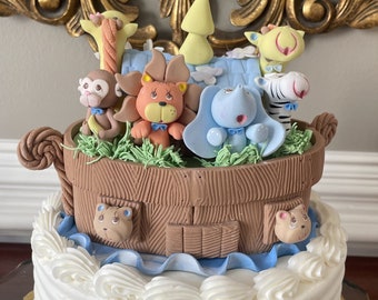 Op de afbeelding: Een witte tweelaagse taart versierd met een bruine fondant ark en verschillende dierenfiguren. De ark is bedekt met een blauw fondant dak en een groene grasbasis. De dieren zijn een leeuw, een olifant, een zebra, een aap en een giraffe. De taart staat op een gouden taartplateau.