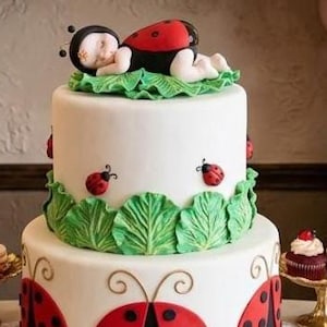 May include: A two-tiered white cake decorated with ladybug designs and green leaf accents. A fondant ladybug figure rests atop the cake. Balloons in red, black, green, gold, and pink create an arch in the background. Additional desserts are on display.