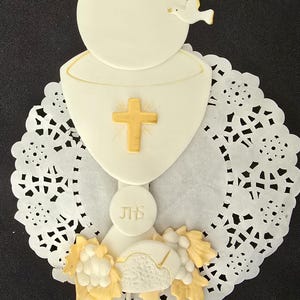 May include: A white communion cake topper featuring a chalice with a gold cross, a dove, and a wafer. The topper is decorated with gold accents and sits on a white paper doily.