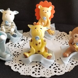 May include: Set of six baby animal cake toppers, each with a blue pacifier. The animals are a zebra, giraffe, lion, monkey, and elephant. Each animal is sitting on a blue blanket.