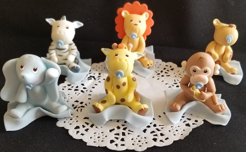 May include: Set of six baby animal cake toppers, each with a blue pacifier. The animals are a zebra, giraffe, lion, monkey, and elephant. Each animal is sitting on a blue blanket.