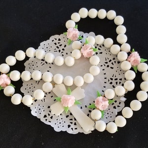 May include: A white beaded rosary with a white cross, decorated with pink flower accents and green leaves. The rosary is presented on a white lace doily against a black backdrop.