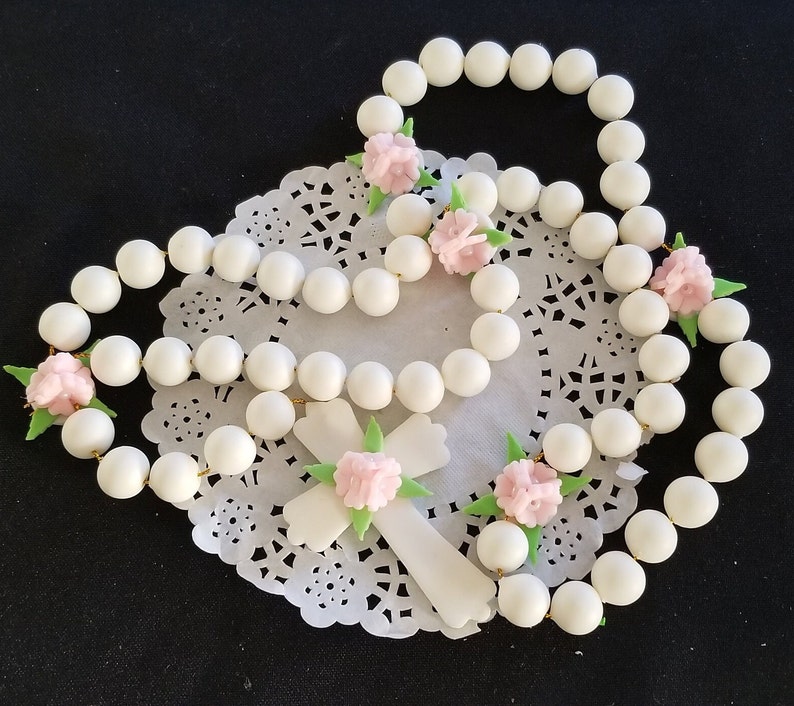 May include: A white beaded rosary with a white cross, decorated with pink flower accents and green leaves. The rosary is presented on a white lace doily against a black backdrop.