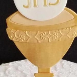 May include: A golden chalice with a white wafer bearing the letters "JHS" in gold. A cluster of purple grapes with green leaves and a small wafer are at the base. The chalice has an ornate design around the rim.