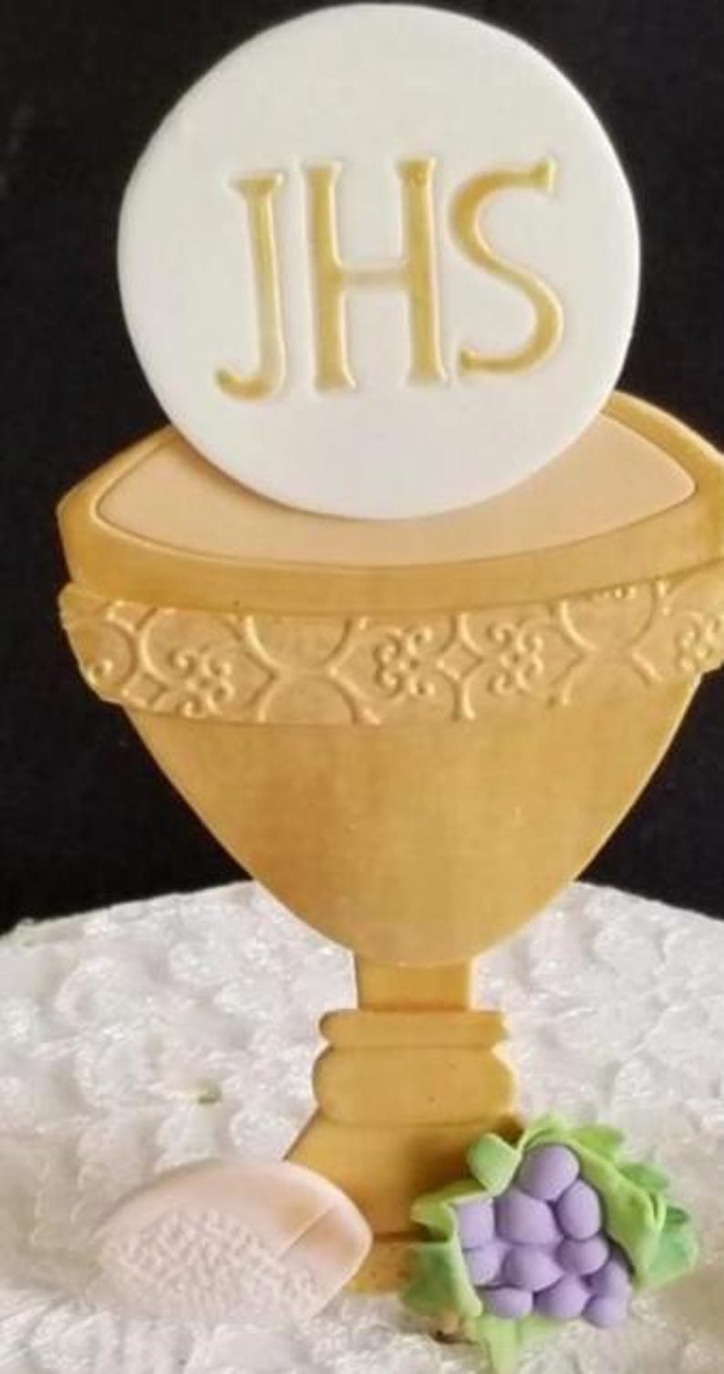 May include: A golden chalice with a white wafer bearing the letters "JHS" in gold. A cluster of purple grapes with green leaves and a small wafer are at the base. The chalice has an ornate design around the rim.