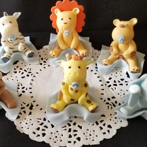 May include: Six baby animal figurines, each sitting on a blue blanket. The animals include a zebra, lion, tiger, giraffe, monkey, and elephant. Each animal is holding a blue pacifier.