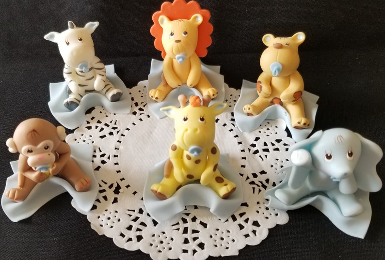 May include: Six baby animal figurines, each sitting on a blue blanket. The animals include a zebra, lion, tiger, giraffe, monkey, and elephant. Each animal is holding a blue pacifier.