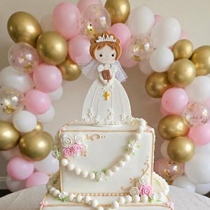 May include: A square white cake decorated with floral details, pearls, and a figurine of a girl in a white dress. The cake is surrounded by a balloon arch in gold, pink, and white. A cross is visible on the figurine.