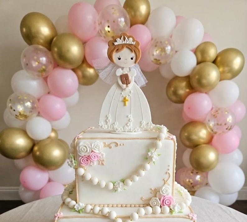 May include: A square white cake decorated with floral details, pearls, and a figurine of a girl in a white dress. The cake is surrounded by a balloon arch in gold, pink, and white. A cross is visible on the figurine.