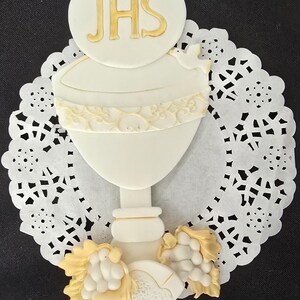 May include: A white communion cake topper featuring a chalice design with gold accents, a dove, and the letters JHS. The topper is set on a white paper doily and includes a cluster of grapes.