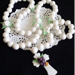 May include: A white rosary with large, round beads and decorative floral accents. The cross pendant features a cluster of purple grapes and a gold detail. The rosary is displayed on a white, lacy doily.
