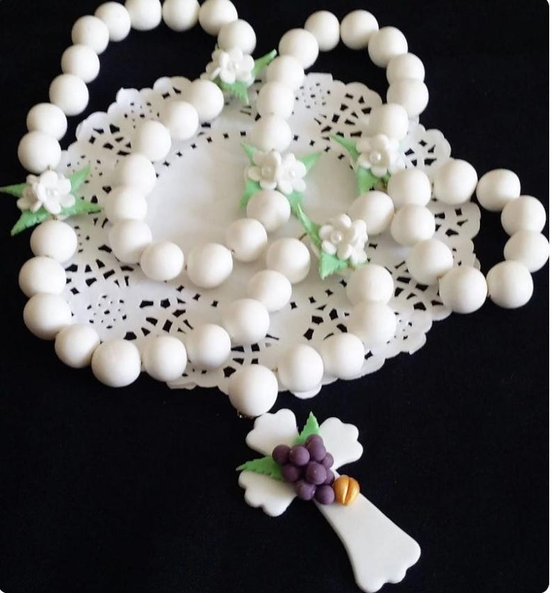 May include: A white rosary with large, round beads and decorative floral accents. The cross pendant features a cluster of purple grapes and a gold detail. The rosary is displayed on a white, lacy doily.