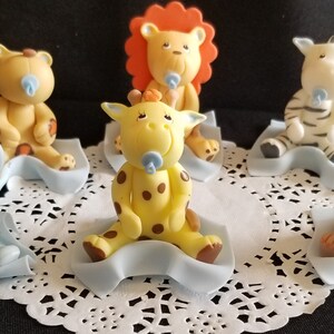 May include: Six baby animal figurines, each sitting on a blue blanket. The animals include a lion, giraffe, elephant, monkey, zebra, and a bear. Each animal is holding a blue pacifier.