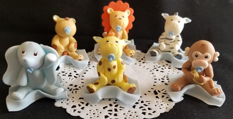 May include: Six baby animal figurines, each sitting on a blue blanket. The animals include a lion, giraffe, elephant, monkey, zebra, and a bear. Each animal is holding a blue pacifier.